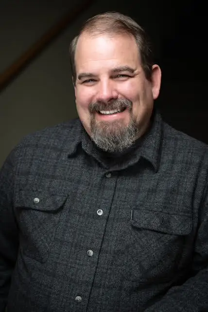 Friendly smiling man with neatly trimmed salt-and-pepper facial hair and short brown hair, dressed in a dark gray button-up shirt, photographed against a softly lit dark background